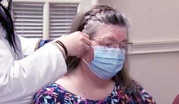 A woman wearing a floral blouse and glasses is seated as a healthcare worker in a white coat checks the fit of her blue surgical mask in a clinical setting with louvered windows.