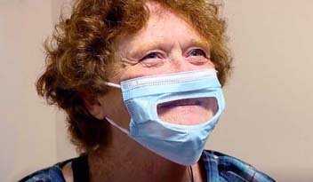 A person with curly red hair wears a blue mask featuring a transparent section revealing their mouth. The person seems to be indoors, wearing a blue plaid shirt.