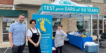 Hearing health awareness event featuring a 'Test your ears at 60 years' booth and informational banner about hearing care benefits.