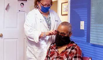 A healthcare worker in a white coat adjusting eyeglasses on a masked person sitting in a clinic room; both are wearing face masks. A poster and framed picture hang on the walls.