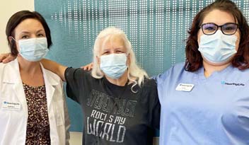 Three women wearing face masks stand with their arms around each other in a hearing care clinic. The woman in the middle has a t-shirt that reads 'Hearing Rocks My World,' and the women on either side wear clinic uniforms.