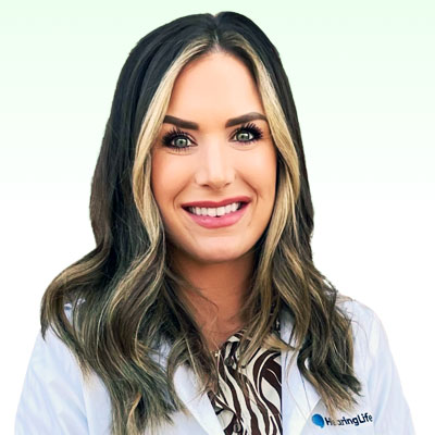 A woman with shoulder-length hair smiles, standing against a pale green background. She wears a white coat with "NursingLife" text on the left side, and a patterned shirt underneath.