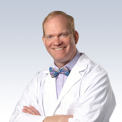 Andrew Brown wearing a white lab coat and a plaid bow tie, standing against a light gradient background.