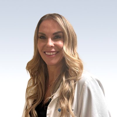A smiling woman with long, wavy blonde hair wears a white lab coat, standing against a plain gradient background transitioning from light gray to white.