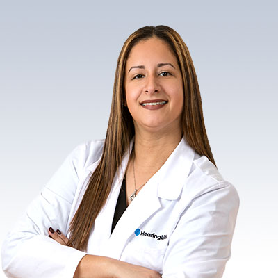 Catherine Rivera wearing a white lab coat with a HearingLife logo, standing confidently with arms crossed.