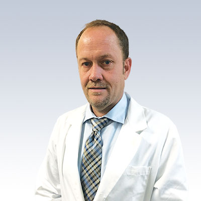 Craig Chaney wearing a white lab coat and a blue checkered tie against a light gradient background.