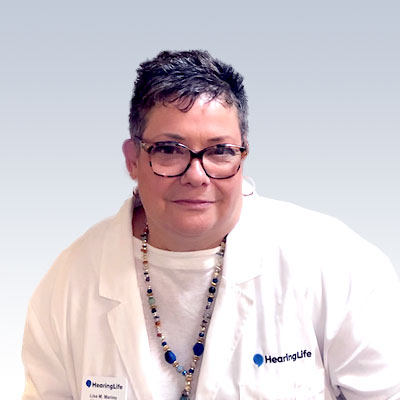 Lisa Marino wearing a white HearingLife coat with a blue and gold beaded necklace against a light gradient background.