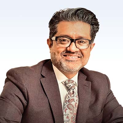 Mario Mendez wearing a brown plaid suit and paisley tie against a light gradient background.