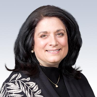 Nancy McGee wearing a black and white patterned shawl, with pearl earrings and a pendant necklace, set against a gradient background.