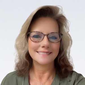 A smiling woman wears glasses and a green top, set against a plain white background, suggesting a professional or friendly setting like a hearing clinic. No text or hearing aid branding is visible.