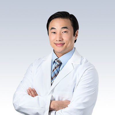 Dan Agrusa in a white lab coat with arms crossed, set against a light gradient background.