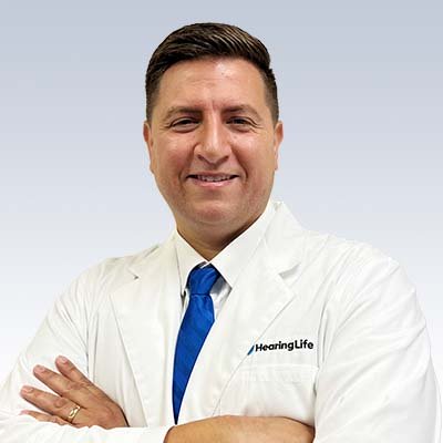 Hearing care professional wearing a white coat with the HearingLife logo and a blue tie, standing confidently with arms crossed.