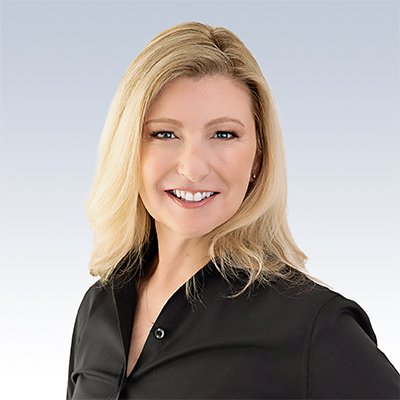 A person with shoulder-length blonde hair, wearing a black collared shirt, is standing against a plain gradient background blending from light blue to white. The face is intentionally blurred. No text or visible branding related to hearing aids, audiologists, or HearingLife is present.