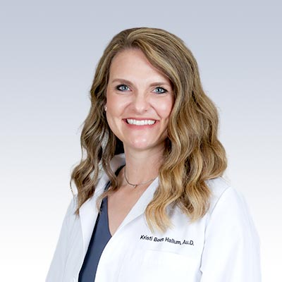 Kristi Been Hallum, Au.D., wearing a white lab coat against a gradient background.