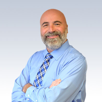 Mark Johnston wearing a blue shirt and plaid tie standing with arms crossed.