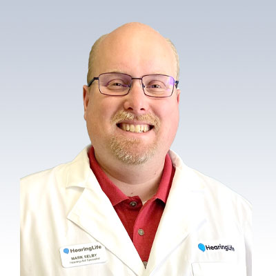 Mark Selby, hearing aid specialist at HearingLife, wearing a white coat with branded name tag.