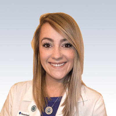 Hearing care professional wearing a white coat with long blonde hair, standing against a light gradient background.