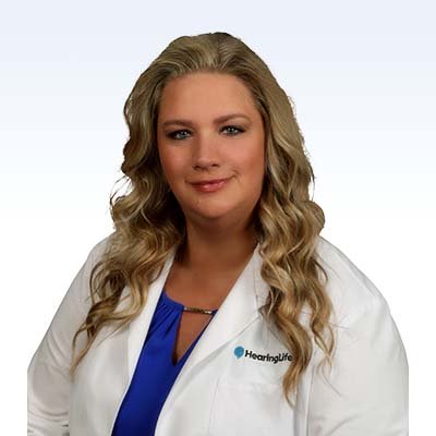Hearing care professional wearing a white coat with the HearingLife logo and a blue blouse, with long wavy blonde hair.