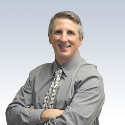 Paul Schneider wearing a grey shirt and patterned tie with arms crossed against a gradient background.