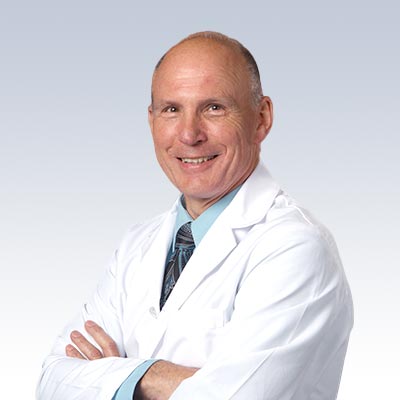 Paul Schuette in a white lab coat with arms crossed against a light gradient background.