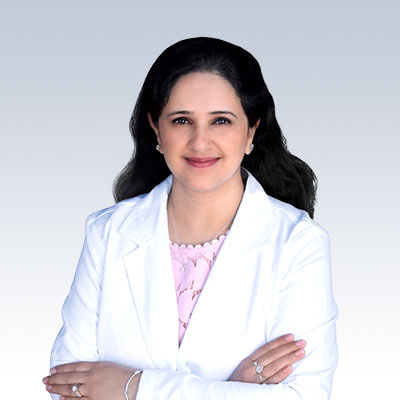 Rashmi Kwatra wearing a white coat with arms crossed against a light gradient background.