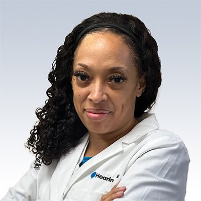Hearing care professional in a white coat with the Hearing logo, arms crossed, on a neutral background.
