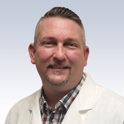 Troy Hill wearing a white coat and plaid shirt, representing a hearing care professional.