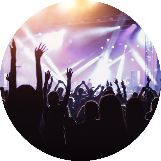 Silhouettes of people raise their arms, dancing and cheering in a packed concert venue with colorful stage lights and spotlights illuminating the background.