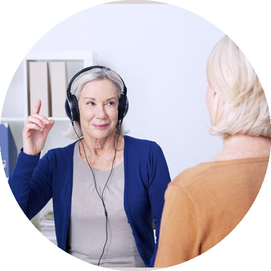 An older woman wearing headphones raises her finger, indicating a hearing test, with another woman seated in front of her in an office setting.