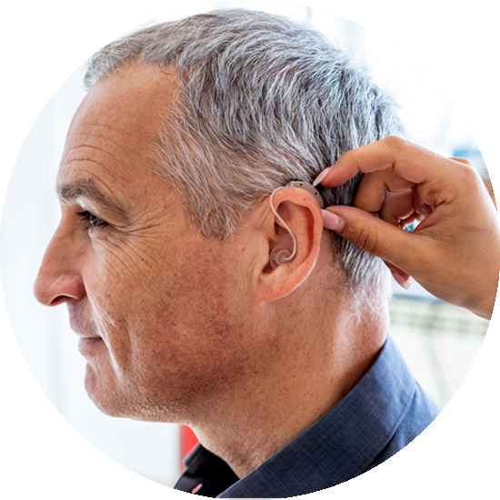 A hand adjusts a hearing aid on an older man’s ear in a bright, indoor setting.