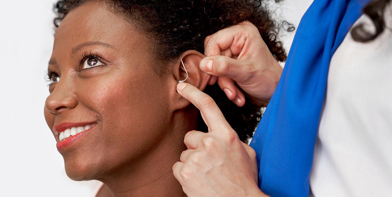 A woman smiles as another person fits a hearing aid into her ear in a well-lit environment, highlighting the process and personalized care involved in adjusting the device.