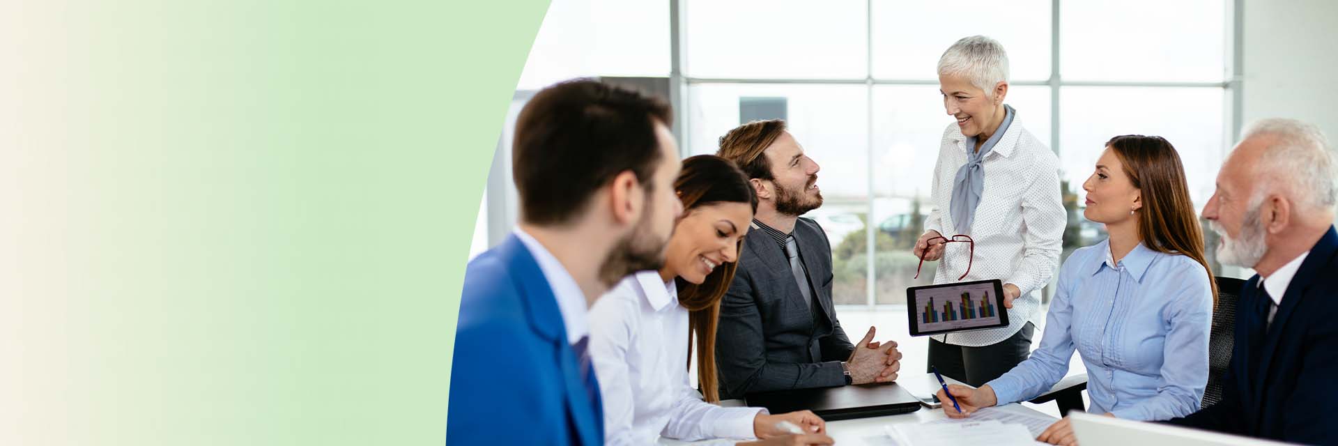 Professionals in business attire engage in a collaborative meeting, discussing data displayed on a tablet and reviewing documents and laptops in a modern office setting.