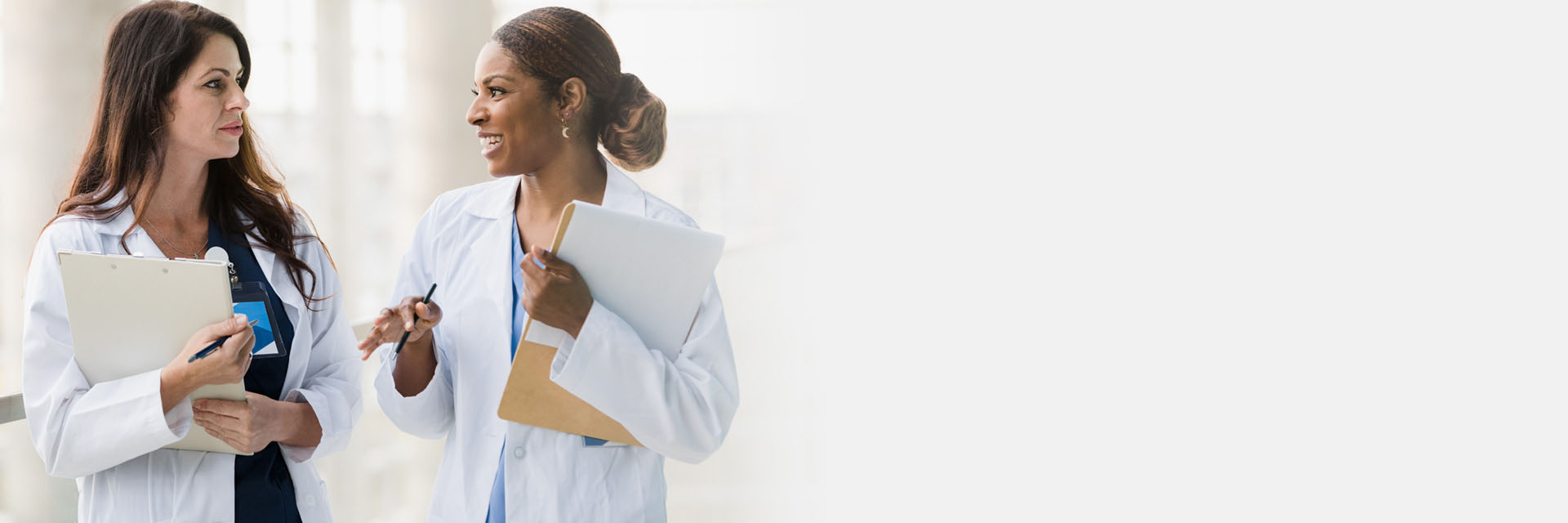 Two professional women in laboratory coats discuss clinical matters in a modern, well-lit hallway while holding clipboards.