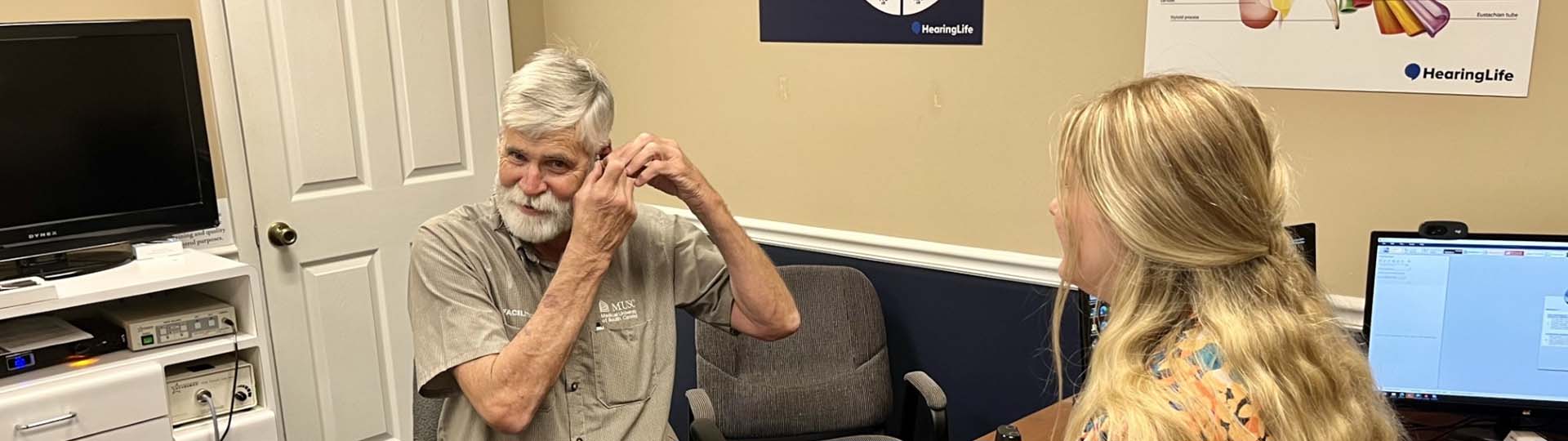 Senior man adjusting hearing aid during consultation with a hearing care professional in an office setting.