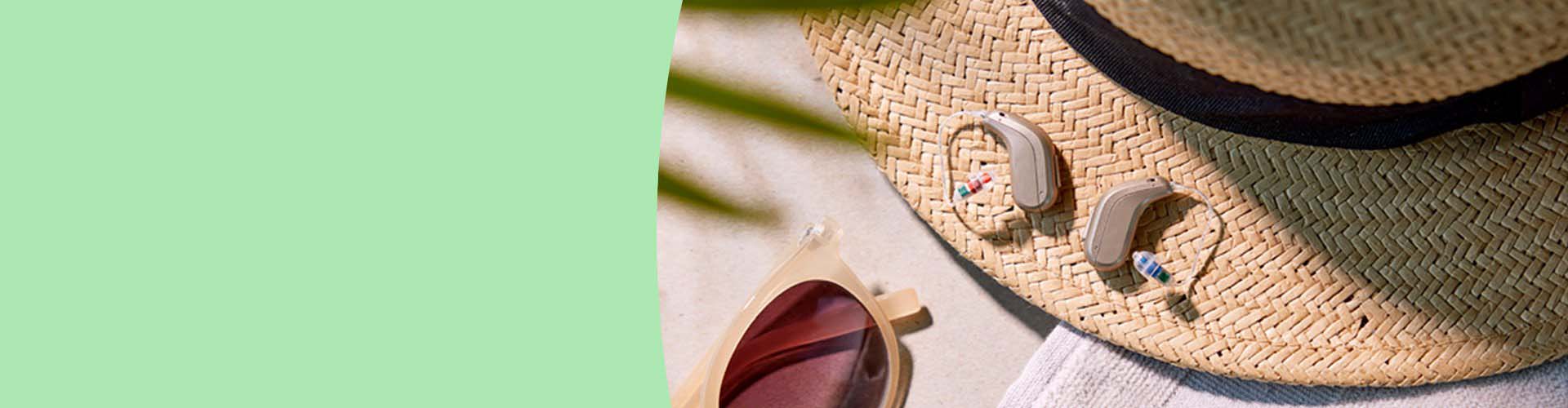 Hearing aids resting on a straw sunhat with beige sunglasses nearby, illuminated by soft daylight.