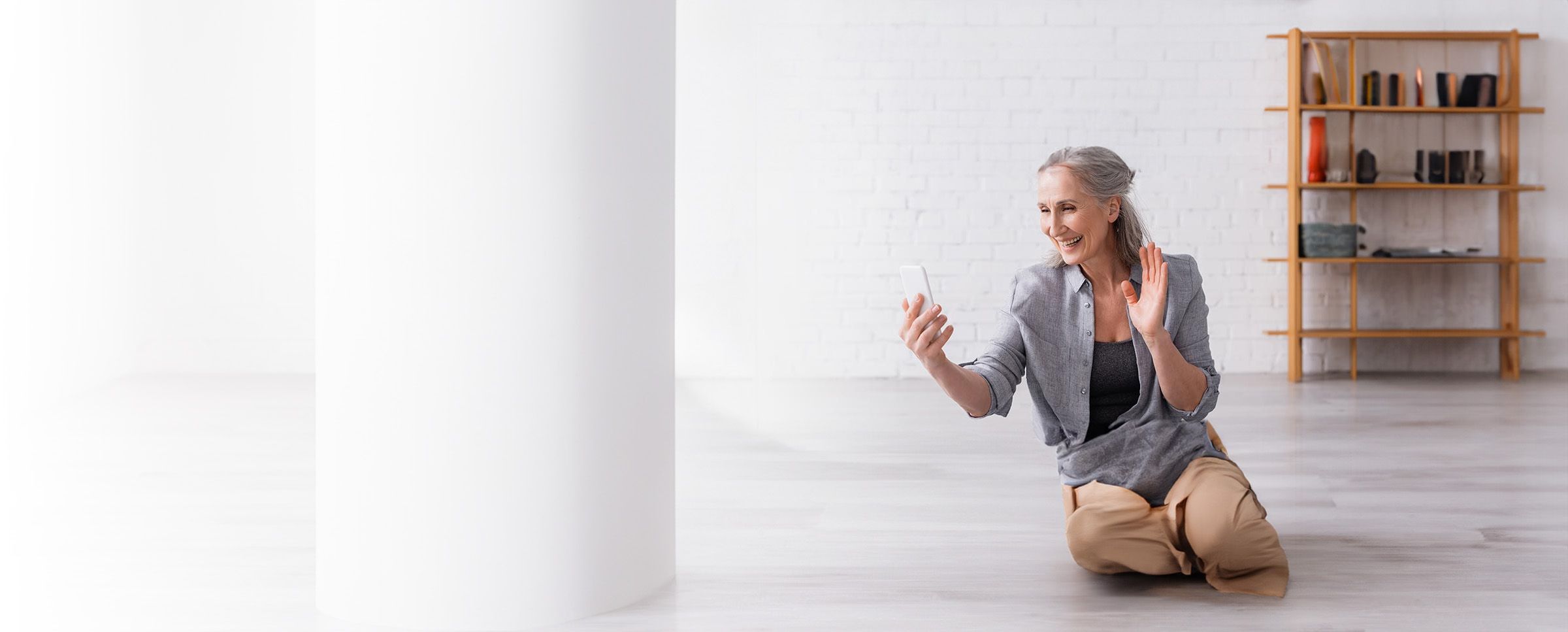 Smiling woman engaging in a video call while sitting on the floor, waving and holding a smartphone.