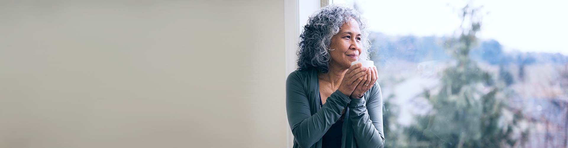 Woman with curly gray hair holds a cup while standing by a window, gazing outside thoughtfully; blurred trees are visible in the background.
