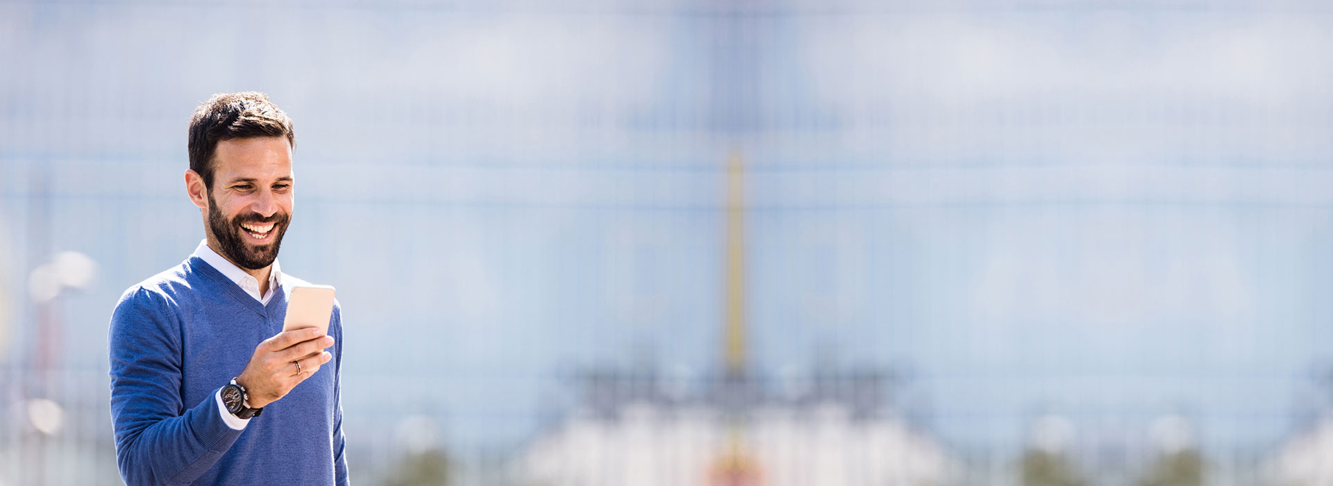 Man smiling while looking at a smartphone, standing outdoors against a blurry urban backdrop.