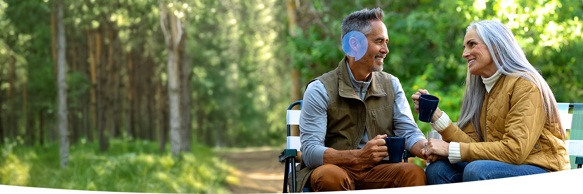 A couple smiling and drinking coffee while camping. A HearingLife speechbubble icon is an overlay on the man's ear.