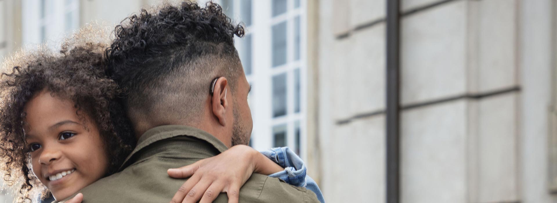 Man wearing a hearing aid embracing a smiling child outdoors.