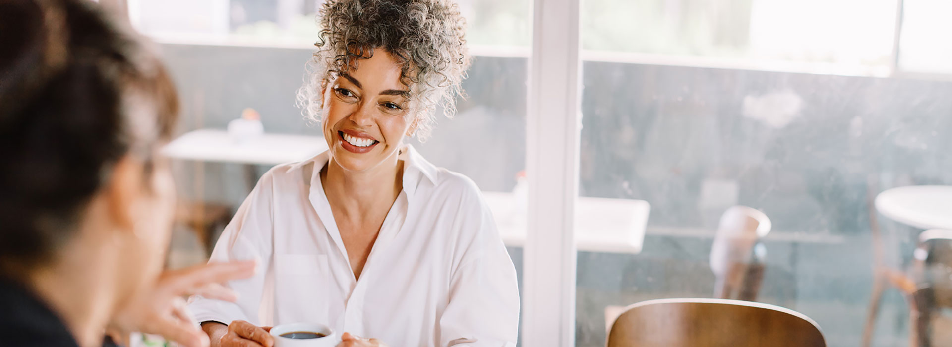 Woman with curly gray hair smiling while holding a coffee cup in a bright restaurant setting.