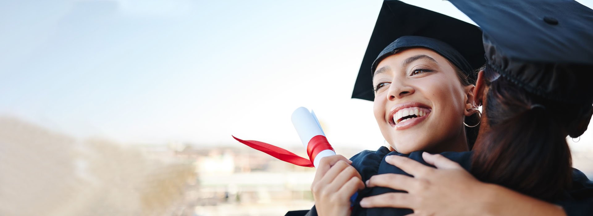 Graduates in caps and gowns celebrating with a congratulatory embrace, holding a diploma tied with a red ribbon.