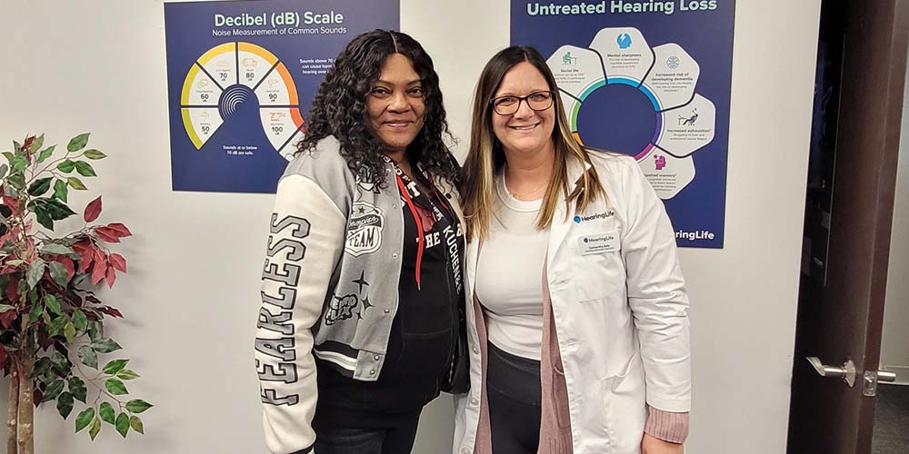 Standing beside a hearing care professional, a woman smiles in front of posters highlighting decibel levels and the impact of untreated hearing loss.