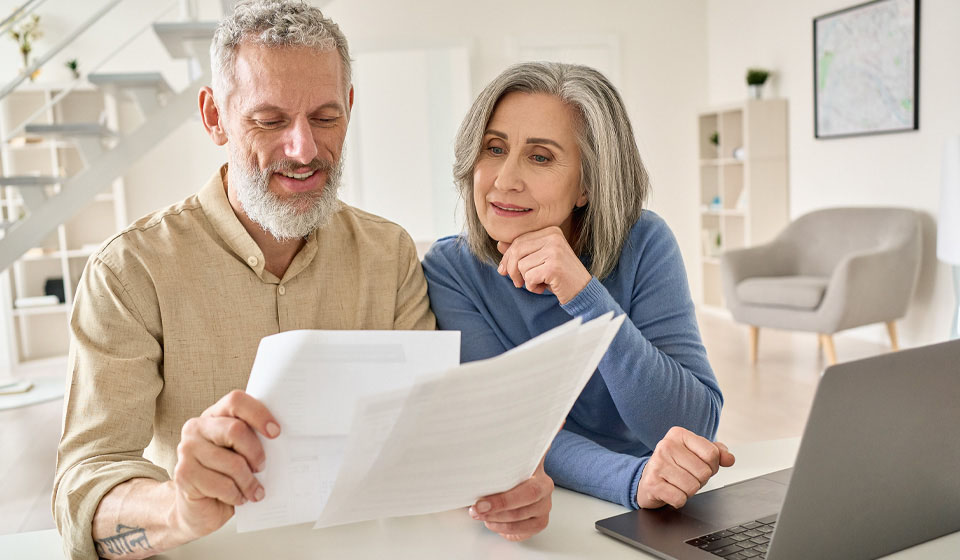 Older couple discussing financial paperwork while seated at a table with a laptop.