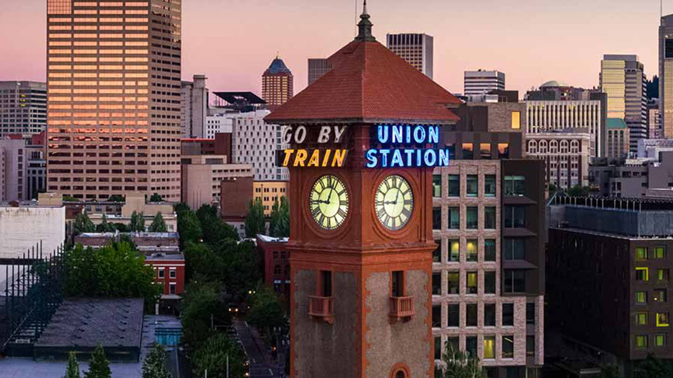 Portland Union Station clock tower with 'Go By Train' neon sign, surrounded by downtown buildings at sunset.
