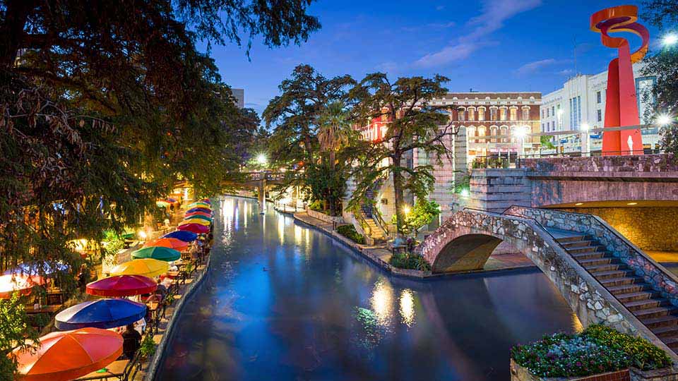 Colorful umbrellas line the San Antonio River Walk at dusk, with vibrant city lights reflecting on the water and iconic architecture in the background.