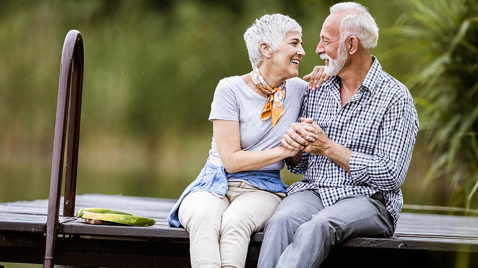 Senior couple smiling and holding hands while sitting together on a wooden dock by a tranquil lake.