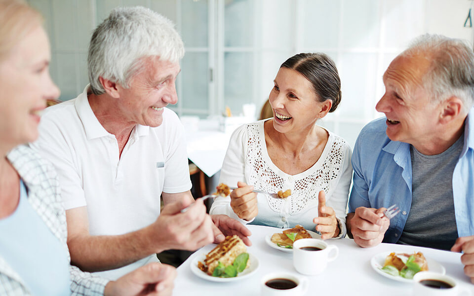 People laughing and talking while sharing a meal at a table with coffee and assorted dishes.