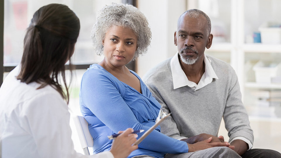 A couple attentively listens while a doctor with a clipboard gestures during a consultation in a bright medical office.