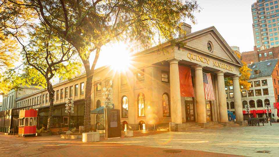 Quincy Market in Boston with sunlight streaming through trees, surrounded by historic buildings and an American flag.